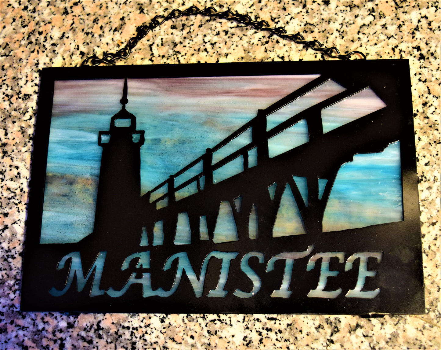 Black custom metal featuring Manistee Michigan lighthouse with Manistee spelled out beneath lighthouse image. With purple and blue beneath metal
