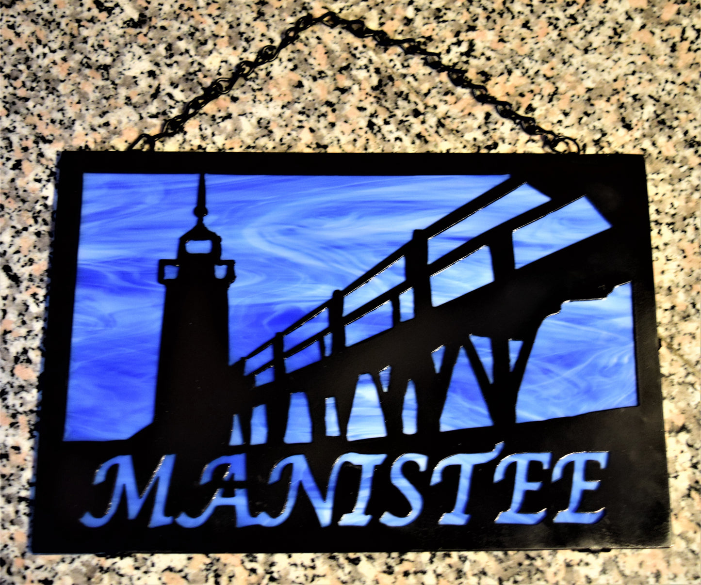 Black custom metal featuring Manistee Michigan lighthouse with Manistee spelled out beneath lighthouse image. With blue background.