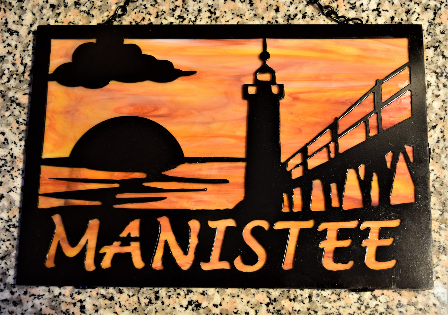 Black custom metal featuring Manistee Michigan lighthouse with cloud and sunset. Manistee spelled out beneath lighthouse image. With Orange and yellow behind metal.