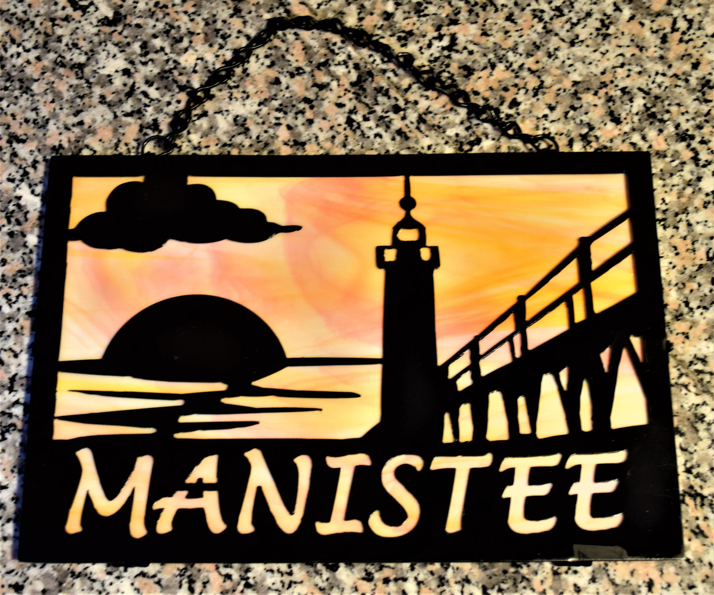 Black custom metal featuring Manistee Michigan lighthouse with cloud and sunset. Manistee spelled out beneath lighthouse image. With Orange and yellow behind metal.
