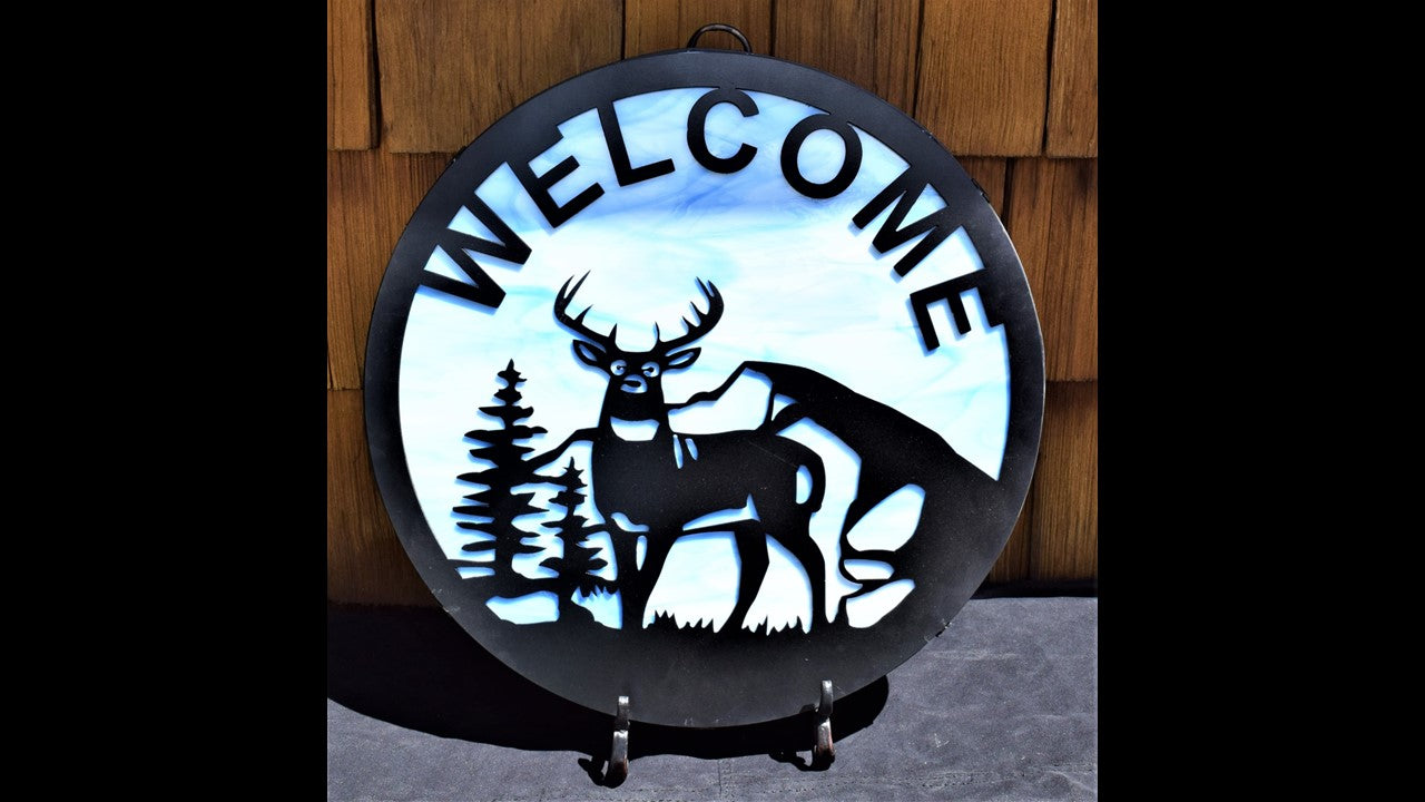 black metal custom welcome silhouette featuring a buck, mountains, trees, and grass with blue stained glass behind silhouette like sky. 