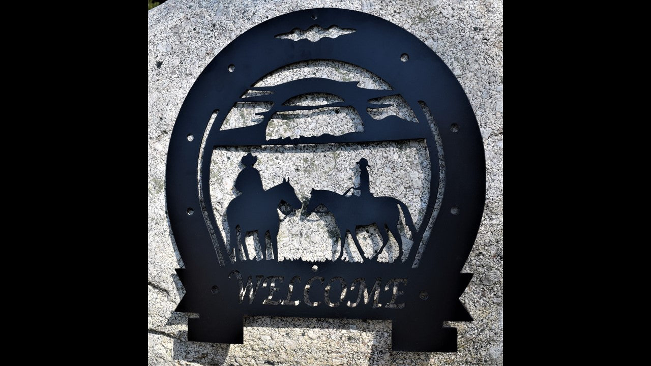 Black custom metal silhouette horse shoe wrapped as boarder featuring a sunset, grass, with a Male riding a horse and a female riding a horse facing each other 
