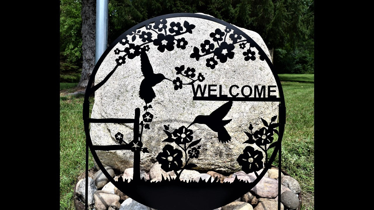 black metal custom welcome silhouette featuring two humming birds and flowers. 24 inches by 24 inches sign stakes are included. Priced at 130 dollars. SKU number YHBW24