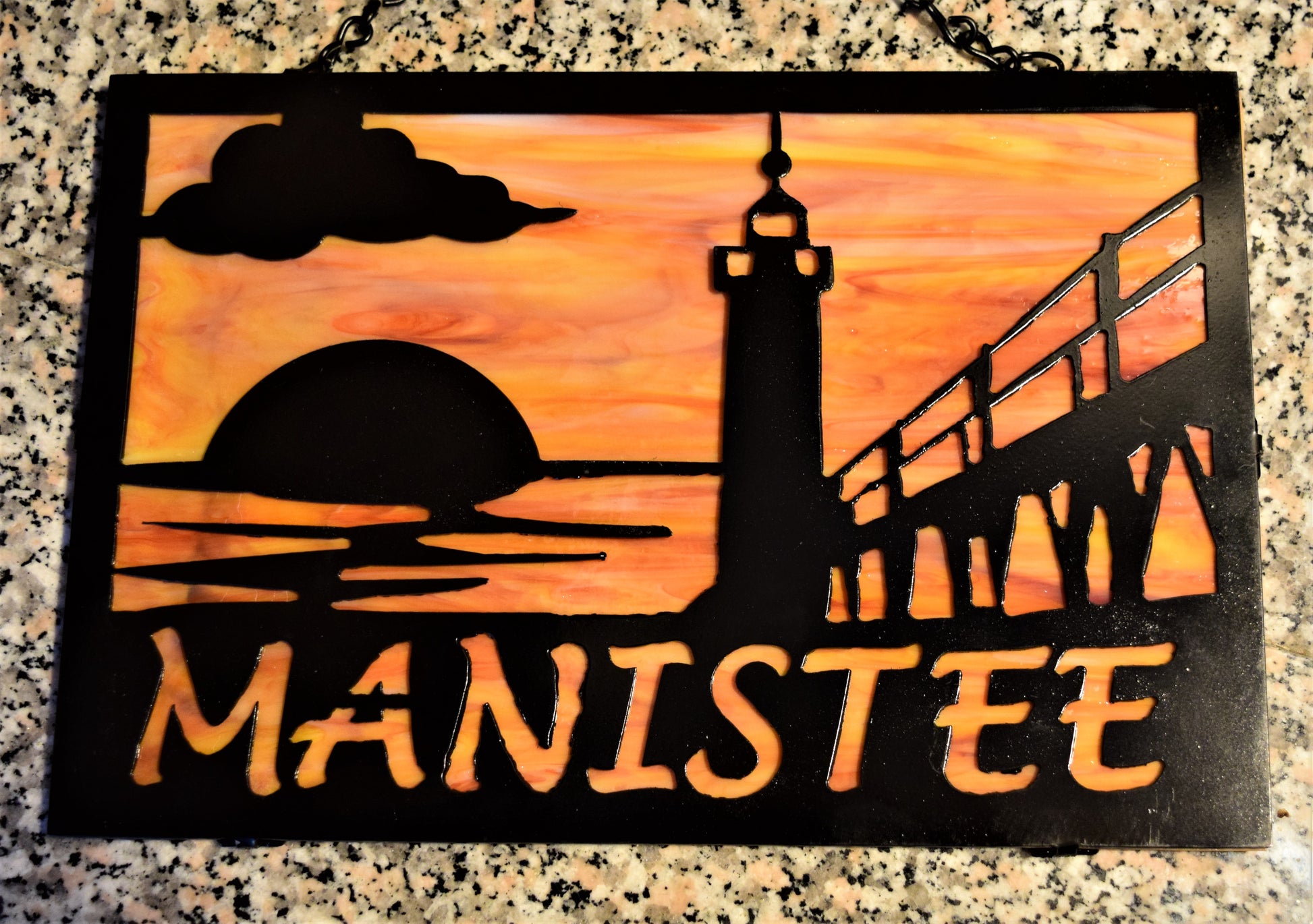 Black custom metal featuring Manistee Michigan lighthouse with cloud and sunset. Manistee spelled out beneath lighthouse image. With Orange and yellow behind metal. 