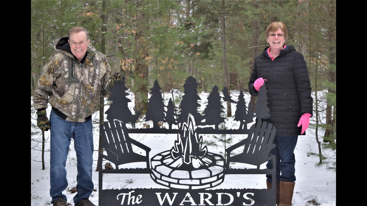 Black custom metal sign featuring the last name The Ward's with a fire pit showing flames, pine tree's, two chairs, a deer and eagle. With customer's standing on each side of their sign. 